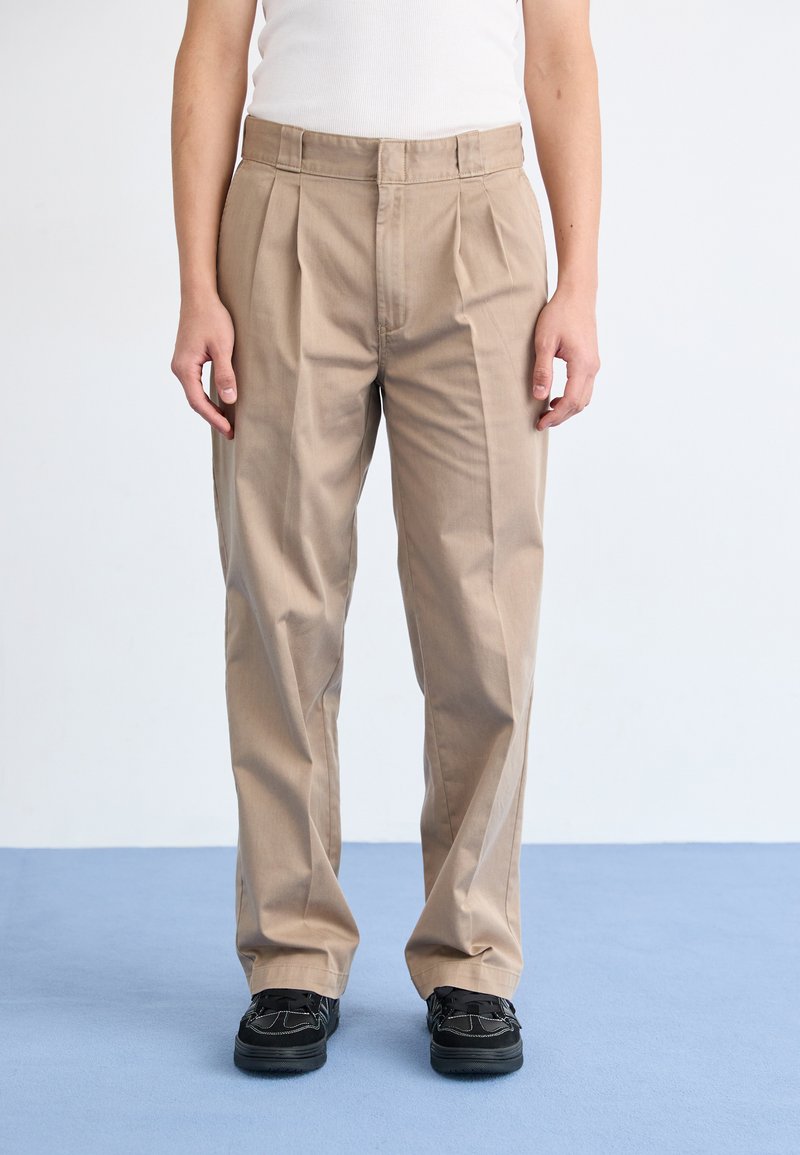 Pantaloni beige plissettati con design a gamba dritta, realizzati in cotone. La cintura è liscia e il tessuto ha una consistenza morbida. Indossati con scarpe nere.
