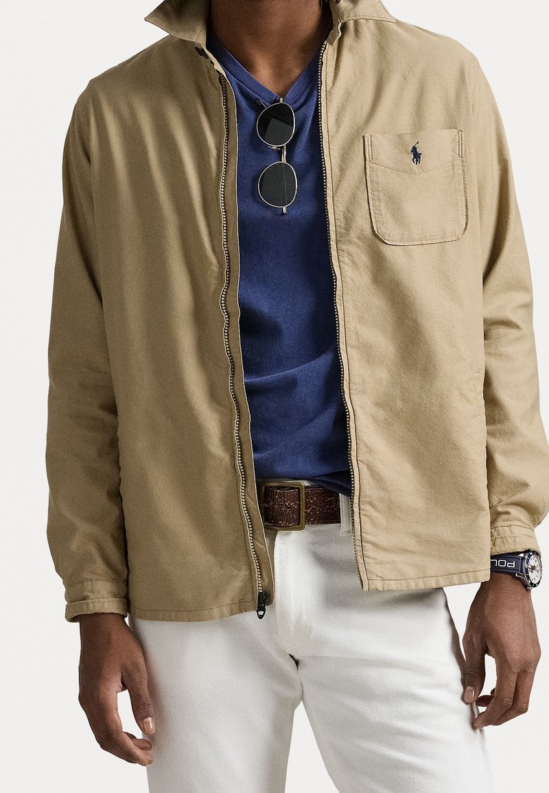 Tan bunda se zipem vpředu, modré tričko pod ní, bílé kalhoty a hnědý opasek. Obsahuje náprsní kapsu a sluneční brýle.