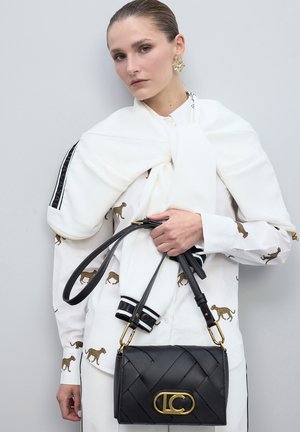 Bolso de cuero negro trenzado con un logo dorado, combinado con una camisa blanca con estampado de leopardo dorado y una bufanda blanca atada al cuello.