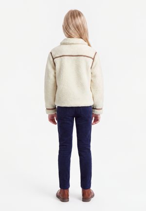 Bambino con lunghi capelli biondi che sta di spalle indossando una giacca sherpa crema, pantaloni blu navy e scarpe marroni su uno sfondo chiaro e uniforme.