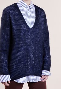 Granatowy sweter z dzianiny z głębokim dekoltem w serek, prążkowaną fakturą i długimi rękawami, nałożony na niebieską koszulę w białe paski z guzikami.