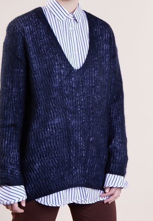 Granatowy sweter z dzianiny z głębokim dekoltem w serek, prążkowaną fakturą i długimi rękawami, nałożony na niebieską koszulę w białe paski z guzikami.