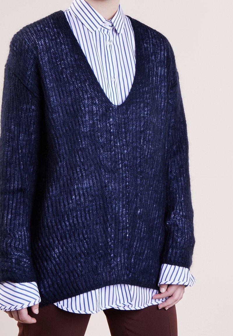 Granatowy sweter z dzianiny z głębokim dekoltem w serek, prążkowaną fakturą i długimi rękawami, nałożony na niebieską koszulę w białe paski z guzikami.