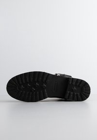 Semelle noire d'une botte robuste avec des crampons profonds et des motifs de traction en forme de croix, positionnée horizontalement sur une surface blanche.