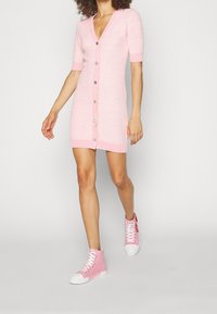 Abito rosa a maglia con maniche corte e scollatura a V, caratterizzato da una chiusura con bottoni sul davanti. Abbinato a sneakers rosa con suola bianca a collo alto.
