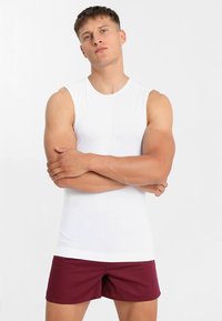 Hvit ermeløs skjorte med tettsittende design, kombinert med mørkerøde shorts. Stoffet ser glatt og lett ut.