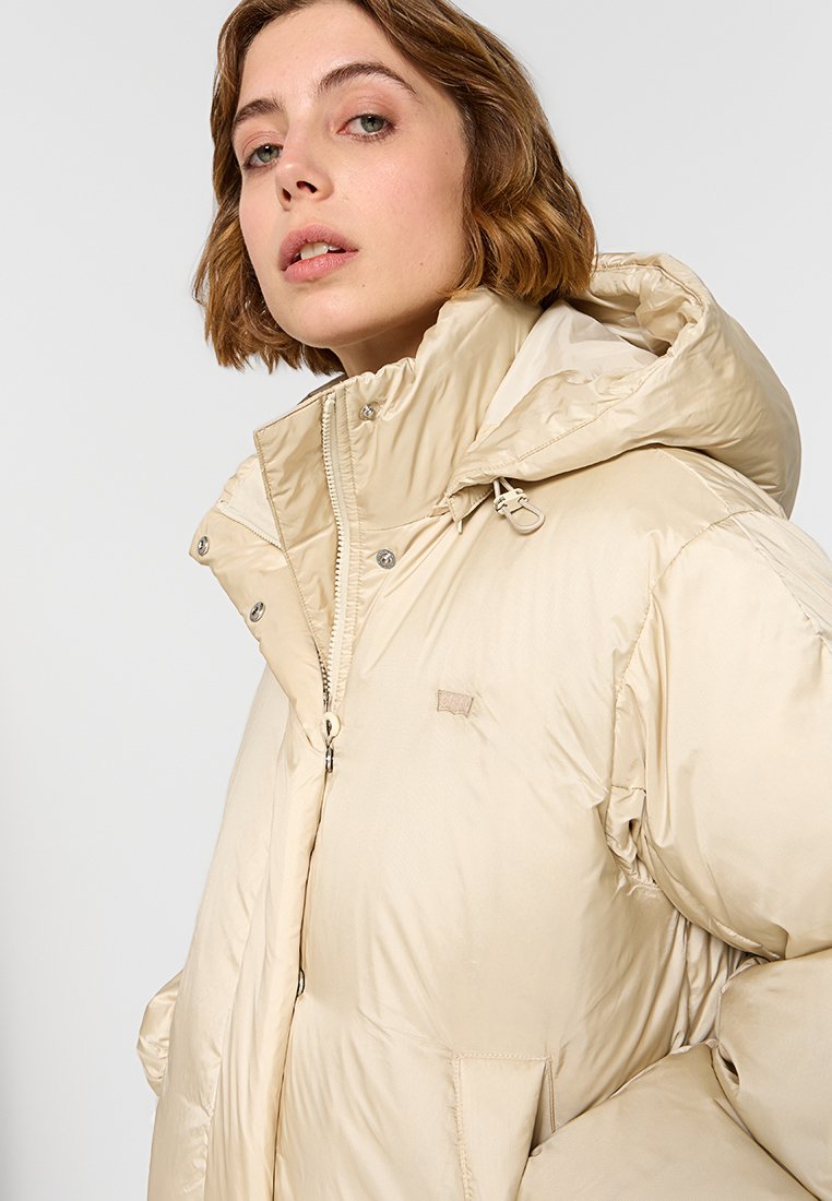 Chaqueta tipo abrigo color beige con capucha, que cuenta con un cuello alto, cierre delantero y botones a presión. Fabricada en un tejido suave y brillante con mínima marca.