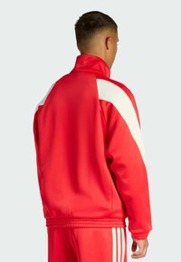 Red athletic jacket with a high collar, white shoulder accents, smooth fabric texture, and fitted cuffs. Rear view showing side stripes.
