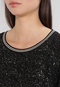 Schwarzer Pullover mit einer strukturierten Stoffoberfläche, die metallische Sprenkler aufweist, und einem gestreiften, gerippten Halsauschnitt in Schwarz und Weiß.