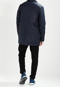 Dunkelblau gepolsterte Jacke mit spitzem Kragen, bestickt mit "Champion" auf dem Rücken, und schwarze Hose mit umgeschlagenen Säumen. Sneakers mit weißen Sohlen.