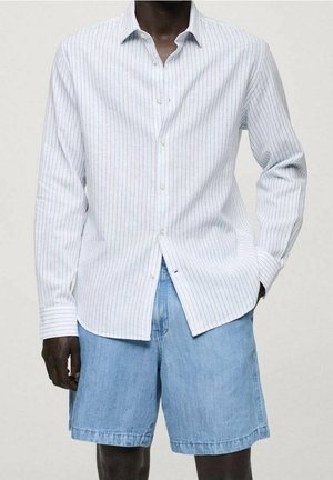 Mand iført en hvid langærmet skjorte med tynde lodrette striber og lyseblå denimshorts mod en ensfarvet baggrund.
