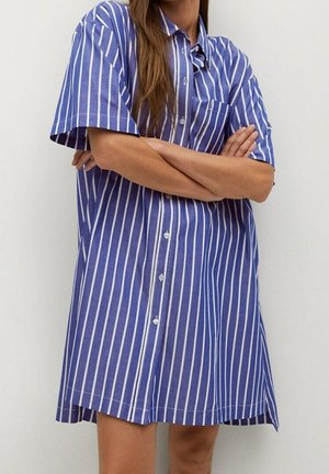 Femme portant une robe chemise à manches courtes rayée verticalement bleu et blanc avec des boutons, debout les bras croisés devant un fond uni.