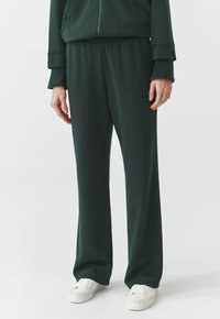 Dunkelgrüne Sweatpants aus weichem Material, mit einem elastischen Bund, lockerer Passform und minimalistischer Designs. Weiße Sneakers werden getragen.