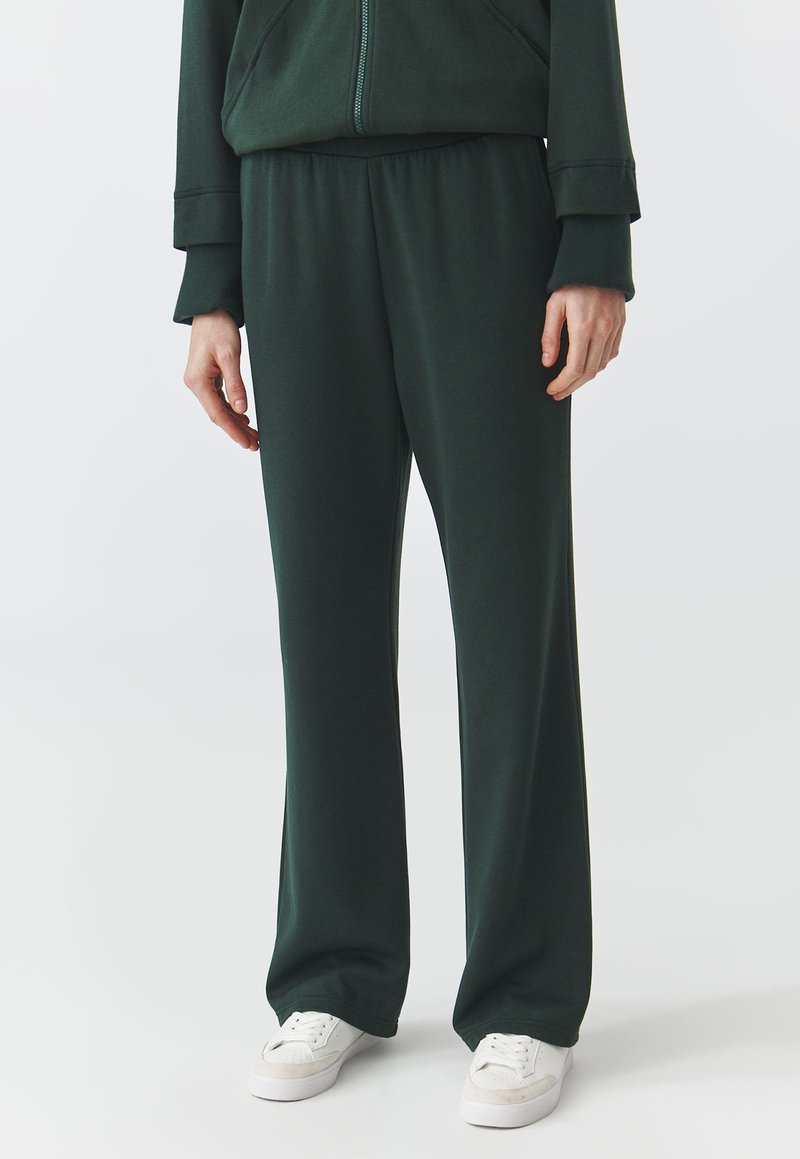 Dunkelgrüne Sweatpants aus weichem Material, mit einem elastischen Bund, lockerer Passform und minimalistischer Designs. Weiße Sneakers werden getragen.