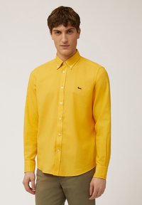 Camisa de botones amarilla de manga larga, hecha de tela tejida, con un pequeño logotipo negro en el lado izquierdo del pecho y botones blancos.