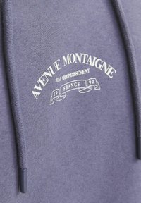 Szürke kapucnis pulóver fehér nyomott logóval, amelyen a következő felirat áll: "Avenue Montaigne 8. kerület 1990 Franciaország." Textúrázott húzózsinórral rendelkezik.