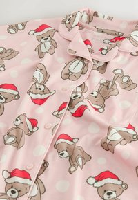 Pink pajama top features a repeated bear pattern wearing red Santa hats, with white polka dots and a button closure. Soft fabric texture.