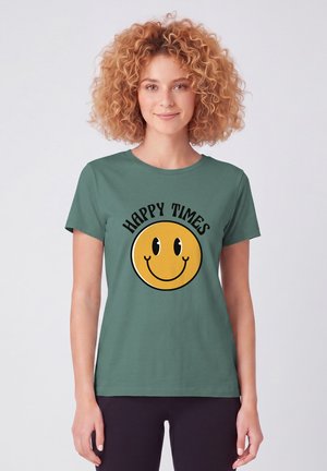 Vrouw met krullend haar die een groen T-shirt draagt met een groot geel glimlachend gezicht en de tekst "HAPPY TIMES" op de voorkant, staand tegen een witte achtergrond.