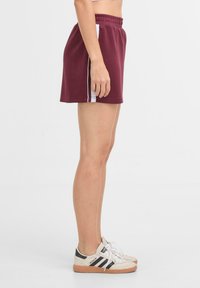 Pantaloni sportivi bordeaux con vita elastica, caratterizzati da strisce laterali bianche. Indossati con scarpe da ginnastica beige e nere. Tessuto dalla texture liscia.