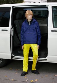 Giacca imbottita blu con tasca frontale, abbinata a pantaloni sportivi giallo brillante con strisce bianche e scarpe nere. Porta del furgone aperta sullo sfondo.