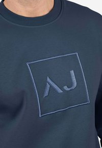 Sudadera azul marino con un cuadrado bordado que lleva las letras "AJ" en un azul más claro. Textura suave y cuello redondo.