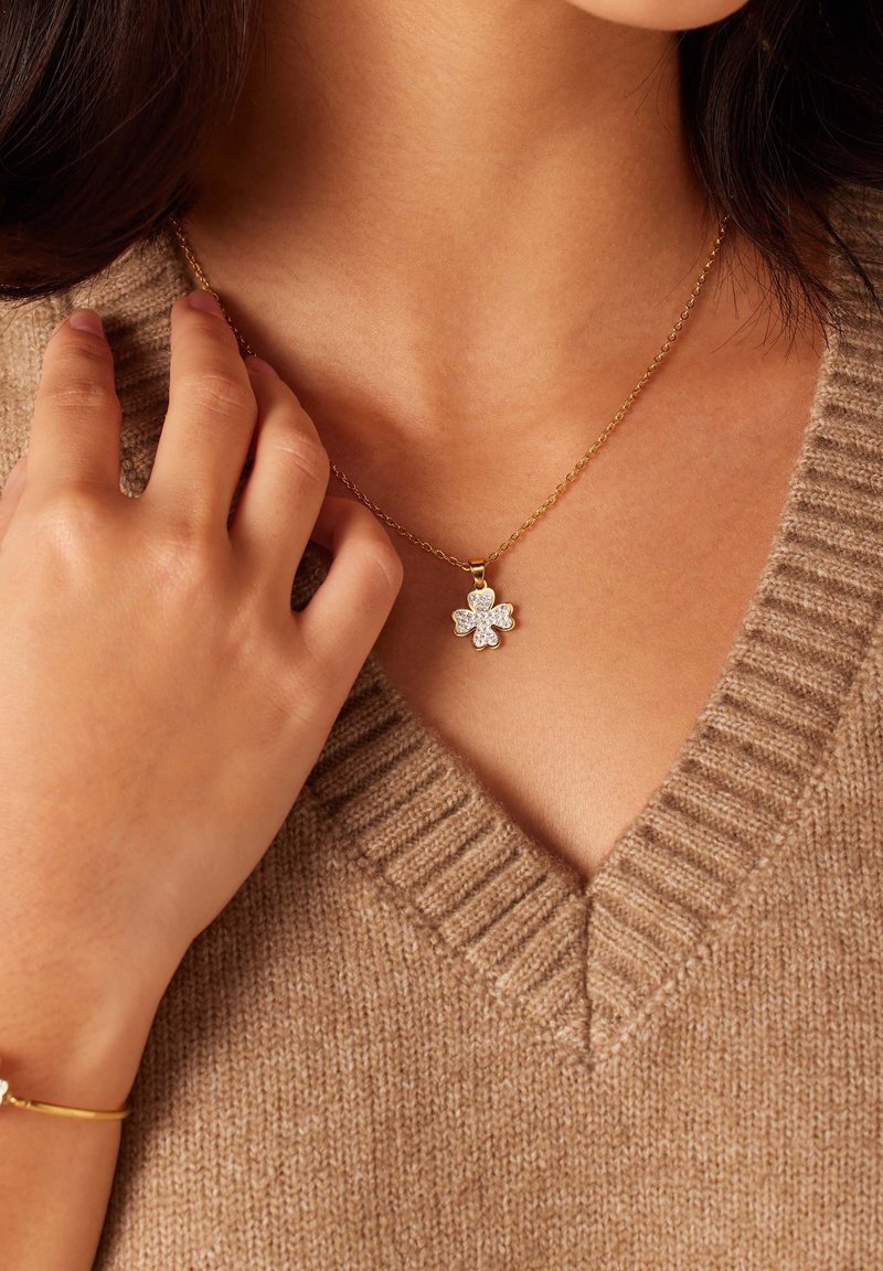 Gouden ketting met een bloemvormige hanger met een cluster van heldere kristallen, gedragen op een beige gebreide v-hals trui.