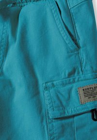 Shorts en tissu turquoise avec une surface texturée, taille élastique, poches latérales et une poche à rabat portant une étiquette avec les détails de la marque.