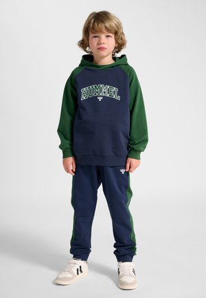 Barn med krøllet blondt hår står foran og vender front med en navy- og grøn Hummel sportshættetrøje og matchende bukser med hvide sneakers.