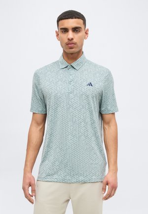 Άνδρας με κοντά μαλλιά φοράει ανοιχτό μπλε πόλο με σχέδιο και μικρό σκοτεινό λογότυπο στο στήθος, στέκεται μπροστά σε απλό φόντο.