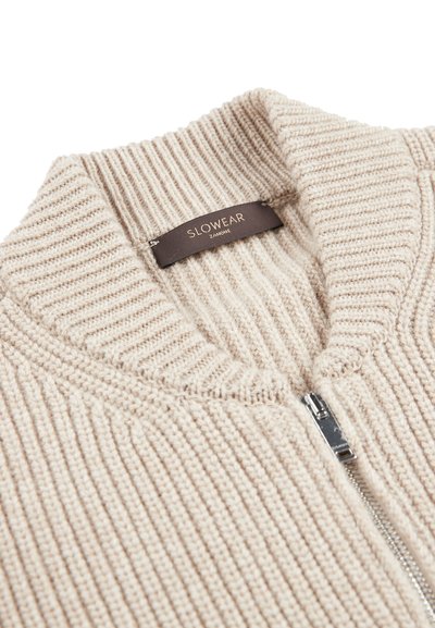 Beige geribde trui met een hoge kraag en een zilveren rits. Voorzien van een stoffen label met de tekst "SLOWEAR". Zachte textuur en aansluitend design.