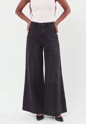 Woman wearing high-waist black wide-leg pants with front pockets, paired with a white sleeveless ribbed top and black pointed heels.