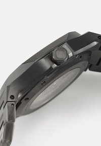 Montre en acier inoxydable noir avec finition mate, couronne octogonale, bords de boîtier texturés et maillons de bracelet lisses. Détails visibles au dos du boîtier.