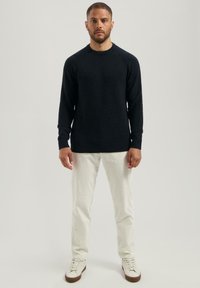 Ein Mann steht frontal mit einem schwarzen Langarmpullover, weißen Hosen und weißen Sneakers mit braunen Sohlen vor einem hellen Hintergrund.