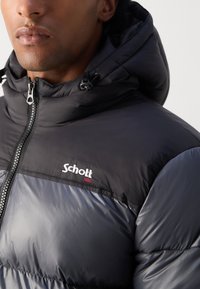 Svart polstret jakke med hette, med glidelås foran og kontrasterende glansfulle og matte teksturer. "Schott" logo på brystet i hvit.