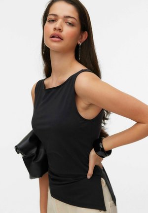Ung kvinde med langt mørkt hår bærer sort ærmeløs top, holder sort clutch og poserer med hånden på hoften mod ensfarvet baggrund.