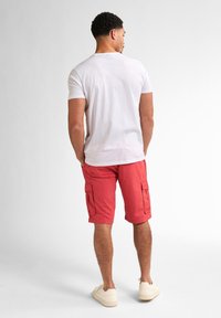 Bermudas rojas con bolsillos laterales, combinadas con una camiseta blanca de cuello redondo. El modelo lleva zapatos de lona beige. Fondo liso.