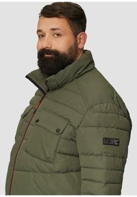 Olivengroene pufferjack met een hoge kraag, voorzien van een rits aan de voorkant, rode accenten en twee borstzakken met drukknoopsluitingen. Logo op de mouw.