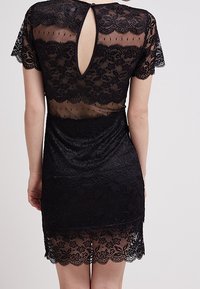 Svart spetsklänning med korta ärmar, genomskinlig design, scallopade kanter, nyckelhålsdetalj i ryggen och blommig spetstruktur längs fållen.