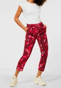 Kvinna klädd i vit ärmlös topp, röd byxor med ormskinnsmönster och beige klumpiga sneakers, stående med händerna i fickorna.