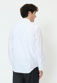 Kiharatukkainen mies selin päin, yll ään valkoinen pitkähihainen paita ja mustat housut, yksinkertaisen valkoisen taustan edessä.