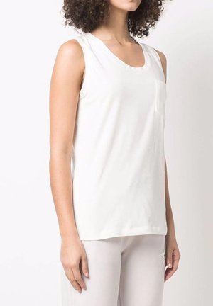 Mujer con pelo rizado, vestida con una blusa de tirantes blanca y pantalones de color claro, de pie frente a un fondo blanco liso.