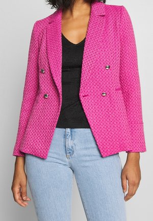 Femme portant un blazer à double boutonnage rose vif texturé par-dessus un haut noir et un jean bleu clair, debout les bras détendus.