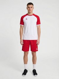 Mladý muž stojí čelem vpřed, oblečený v bílém a červeném sportovním tričku s krátkým rukávem, červených šortkách, bílých ponožkách a černých sportovních botách na jednobarevném pozadí.
