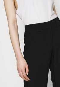 Černé kalhoty na míru vyrobené z hladké látky, s vysokým pasem, poutek na opasek a bočními kapsami. V kombinaci s bílým topem bez rukávů.