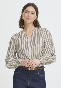 Blouse rayée beige et marine avec un col en V, des manches bouffantes et des poignets froncés. Portée avec un denim foncé et une ceinture marron avec une boucle dorée.
