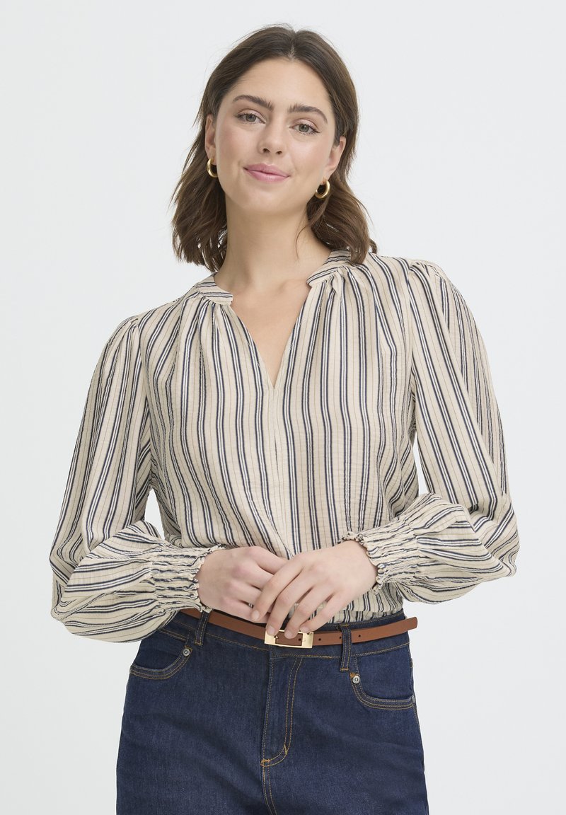 Blouse rayée beige et marine avec un col en V, des manches bouffantes et des poignets froncés. Portée avec un denim foncé et une ceinture marron avec une boucle dorée.