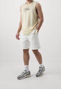 Lichtgele mouwloze sporttop, witte shorts en grijze sportschoenen met accenten. Bevat een mesh-textuur en logo-ontwerp op de top.