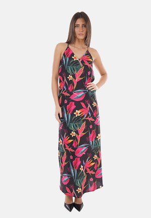 Donna in piedi che indossa un abito lungo nero senza maniche con vivaci motivi floreali tropicali rosa, arancioni e verdi e scarpe nere con punta.