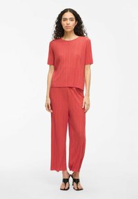 Top rosso plissettato con maniche corte, abbinato a pantaloni ampi plissettati. Texture liscia, design semplice, completato da sandali neri.