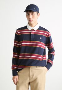 Marineblå og burgunderstripet langermet polo med hvit krage og brodert logo, kombinert med khakibukser og en marineblå caps.